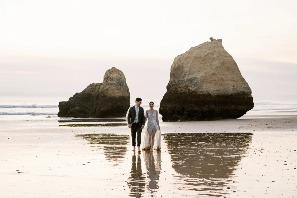 A newlywed couple walks barefoot along the shoreline at sunset, with towering rock formations reflecting in the wet sand.