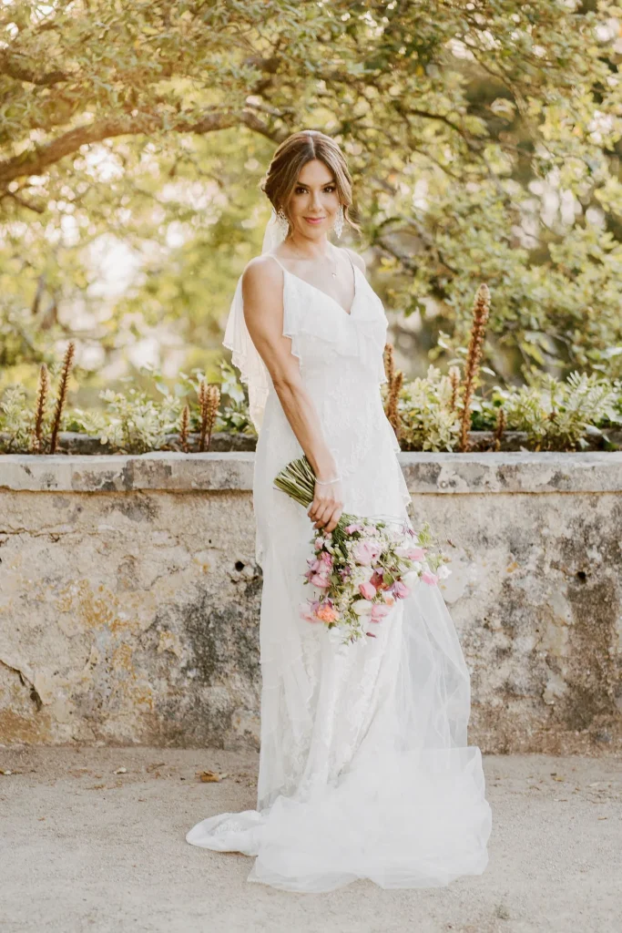 A bride in an elegant lace gown holding a pastel-colored bouquet, standing against a stone wall surrounded by greenery.