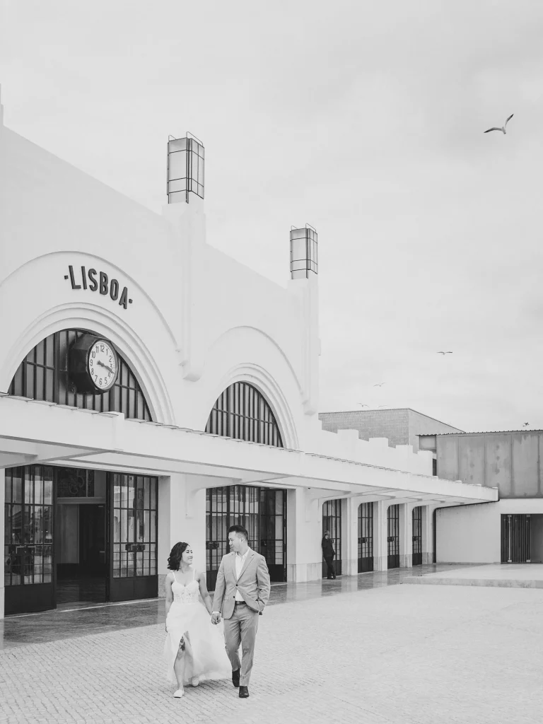 A black-and-white photograph of a couple walking hand-in-hand in front of a historic train station in Lisbon, Portugal.
