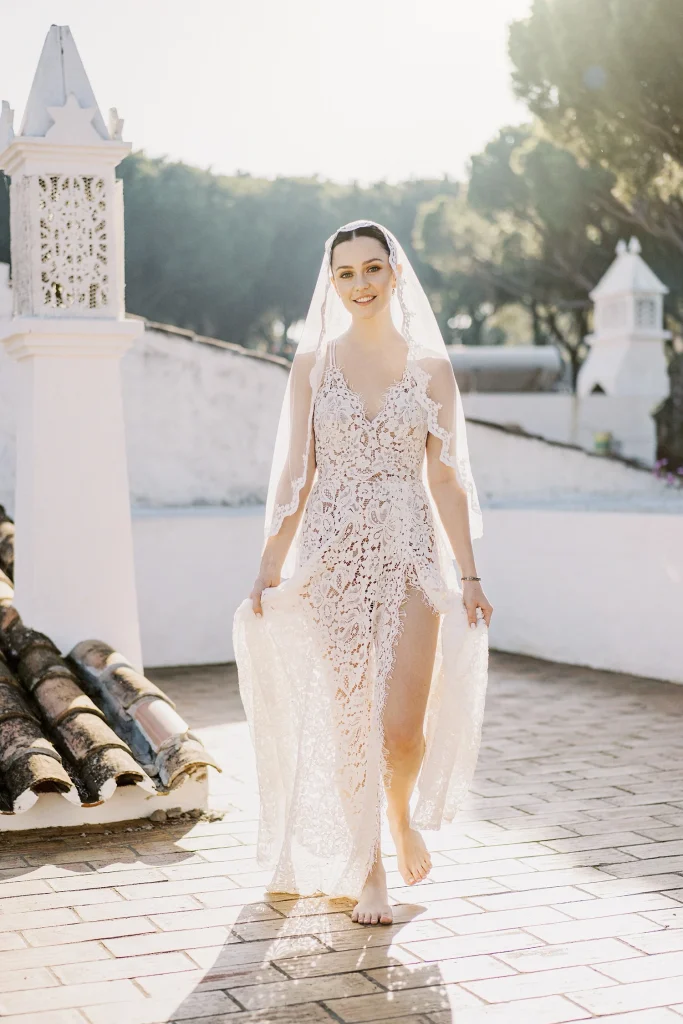 A bride in an intricately designed gown with a long veil walks confidently through a sunlit courtyard.