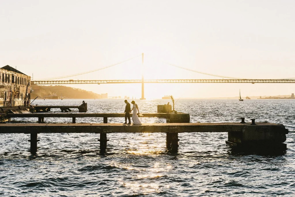 Silhouettes of a couple standing on a wooden pier, framed by the setting sun and a suspension bridge in the distance.