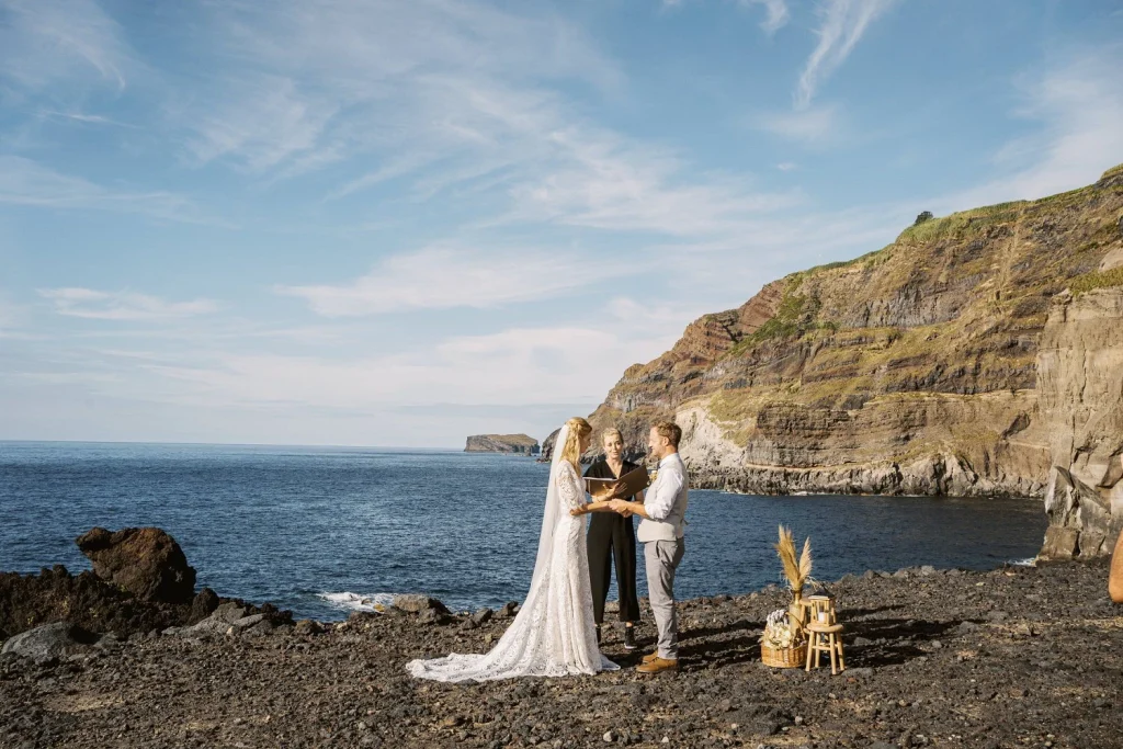 A couple exchanges vows on a rocky shoreline with towering cliffs in the background, under a bright blue sky.