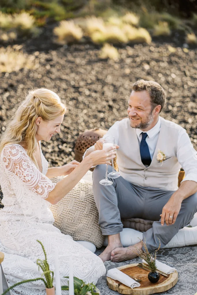 A newlywed couple enjoys a relaxed outdoor picnic, clinking champagne glasses and sharing a joyful moment on a bohemian-style setup with cozy cushions and rustic decor.
