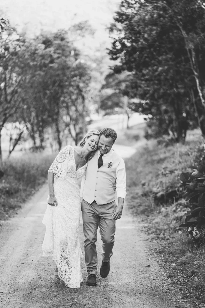 A black-and-white portrait of a bride and groom walking down a country path, leaning into each other with relaxed smiles.