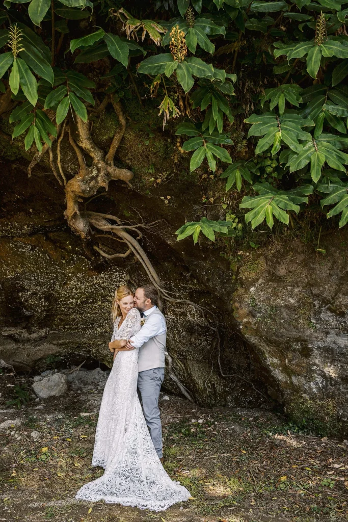 A couple embraces under the sprawling roots of an ancient tree, surrounded by lush greenery.