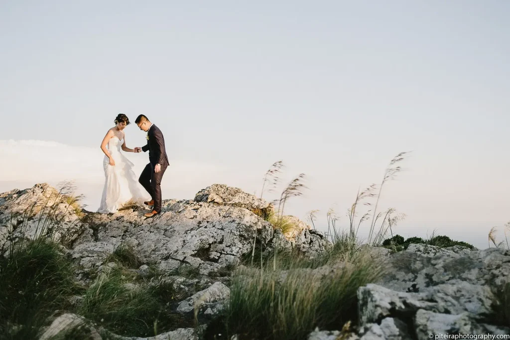 Bride and groom standing atop a scenic rocky hill, sharing a quiet moment surrounded by nature.