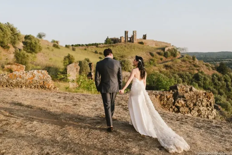 Bride and groom walking hand in hand towards historic castle ruins, surrounded by rolling green hills and golden sunlight.