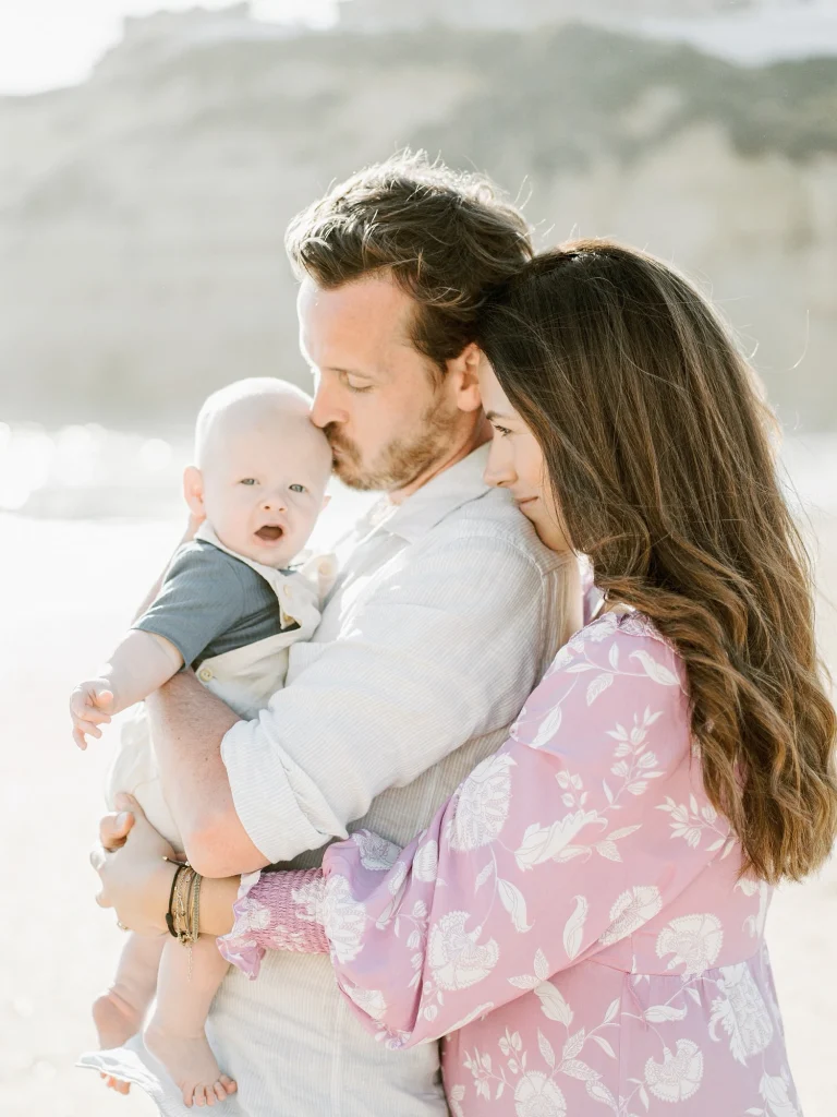 A young couple embraces their baby outdoors, with the father kissing the infant’s head while the mother holds them close.