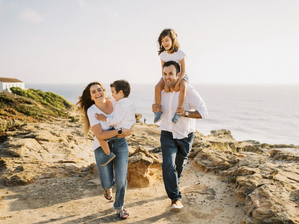 A joyful family adventure by the coast, with a father carrying his daughter on his shoulders while the mother and son walk beside him.
