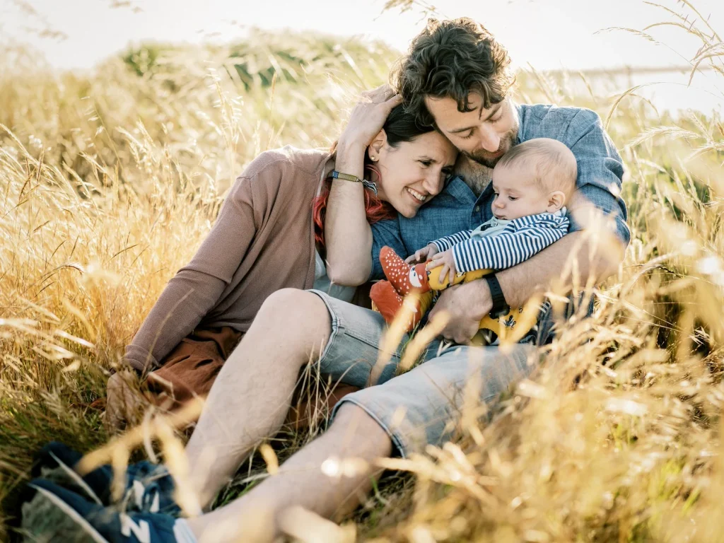 A young couple sits in a sunlit field, embracing their baby while surrounded by golden grass.