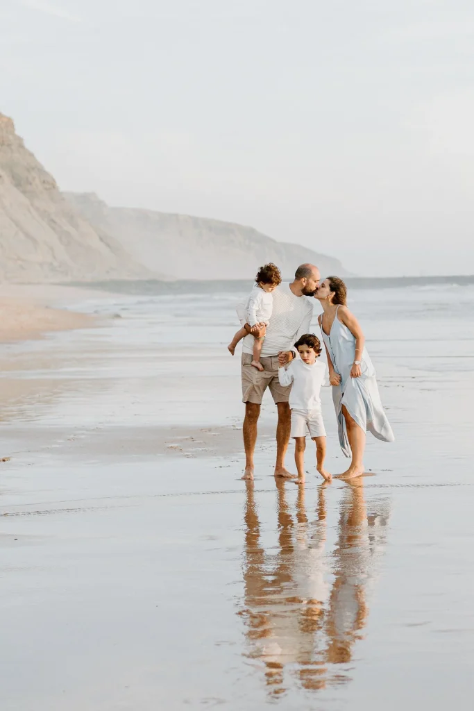Family of four walking along the shoreline, children laughing as waves touch their feet.