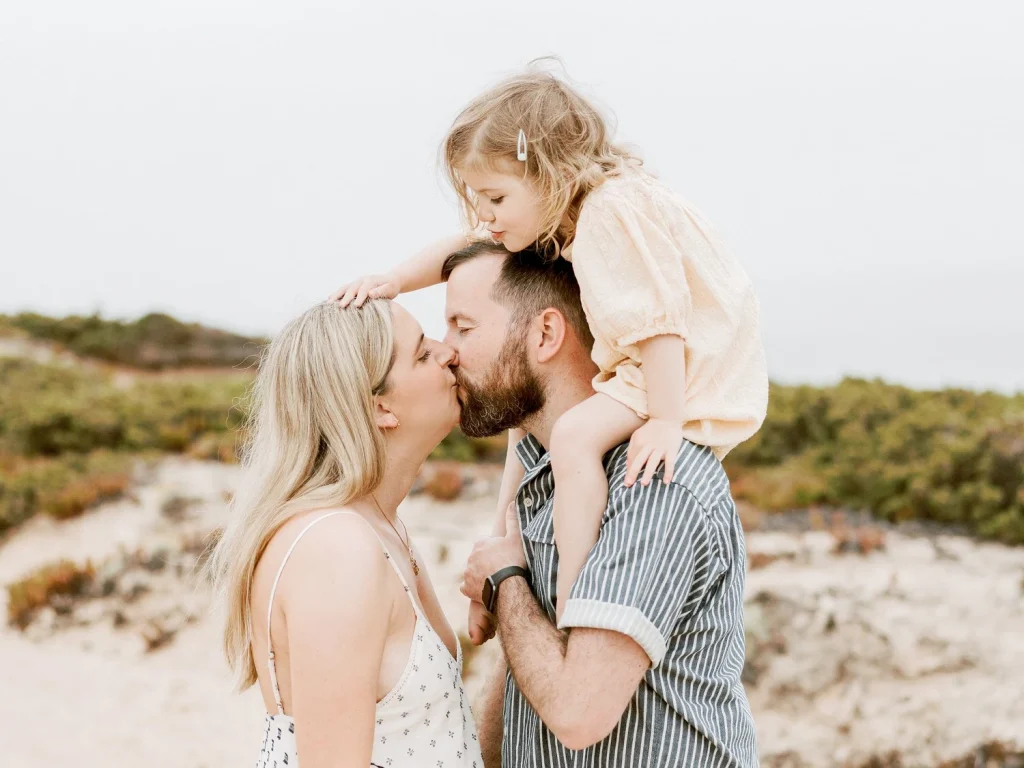 Mother and father sharing a kiss while their young daughter sits on the father’s shoulders, smiling down at them.