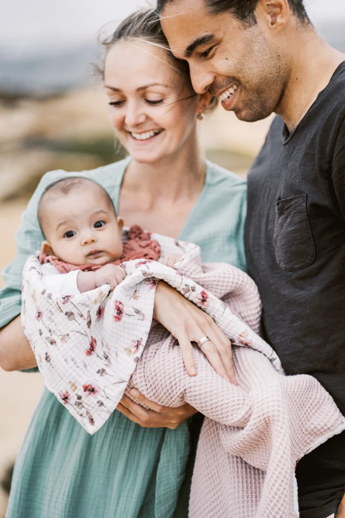 Close-up of parents holding their baby wrapped in a cozy pink blanket, smiling down at their child.