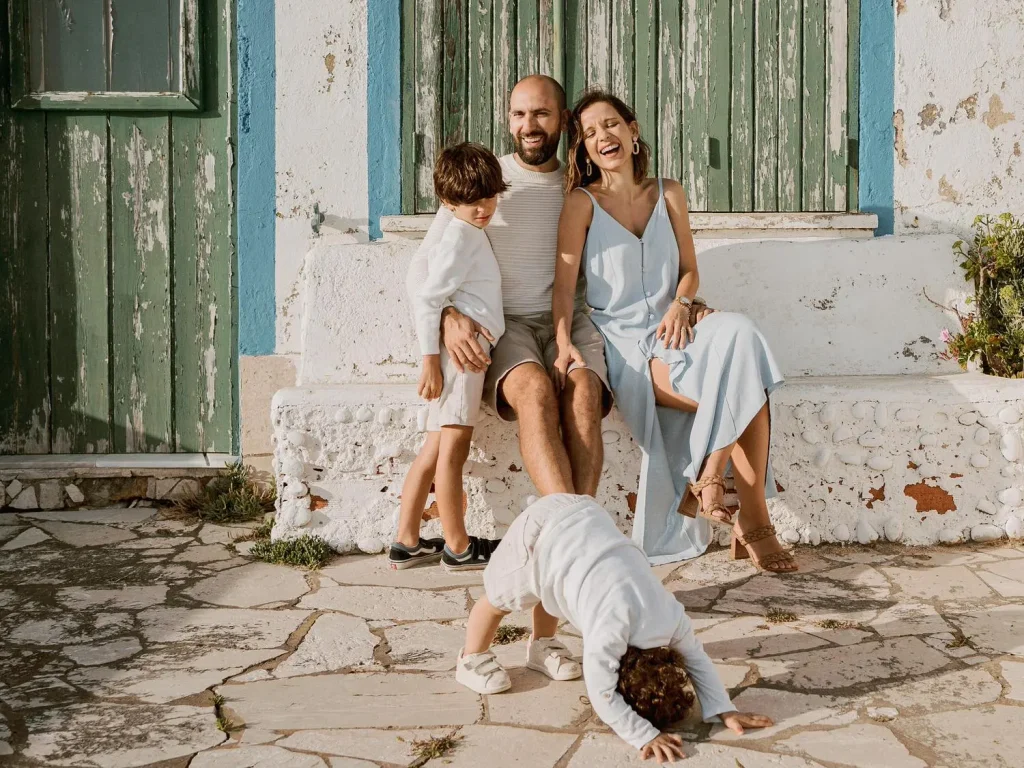 Playful family sitting on a rustic bench outside a weathered blue-green building, with children laughing and playing around them.