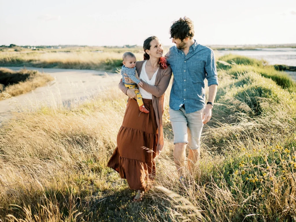 Smiling family walking through a sunlit field, with the mother holding a baby and the father by her side.