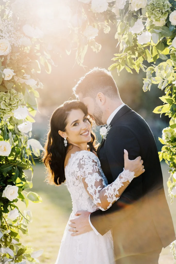 A bride in a lace wedding dress smiles at the camera while embracing her groom under golden sunlight.