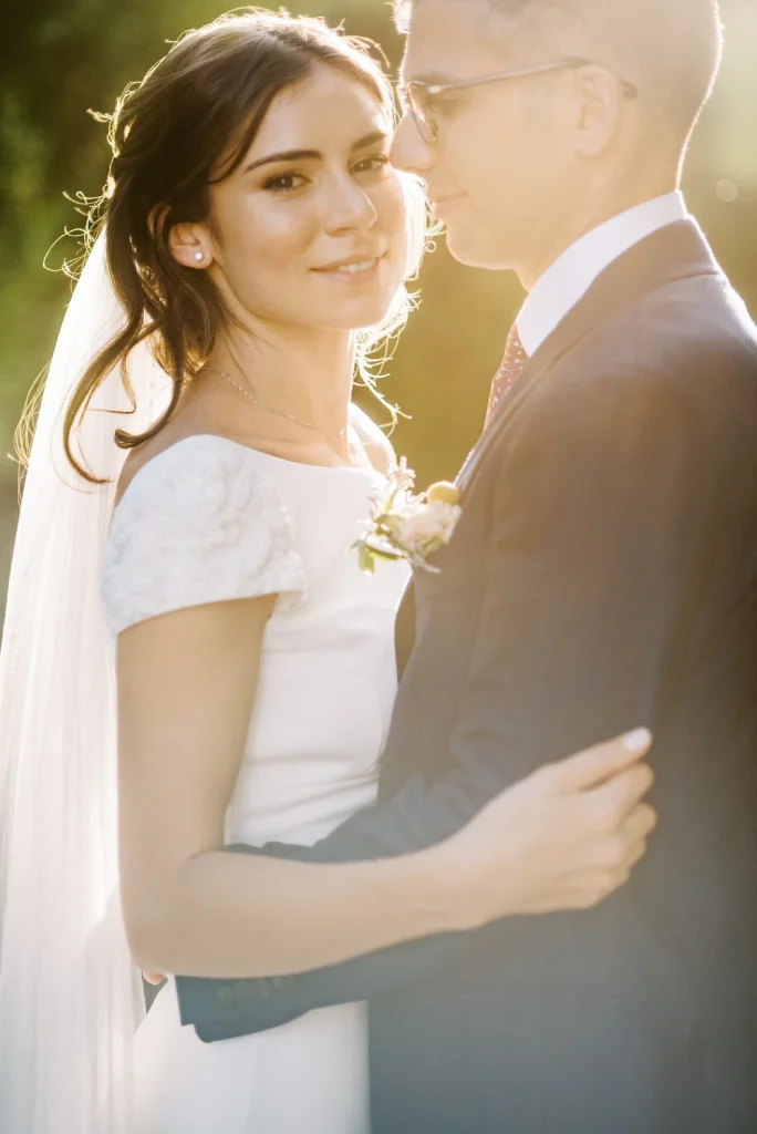 Bride and groom embracing on a sunlit garden path, surrounded by lush greenery.