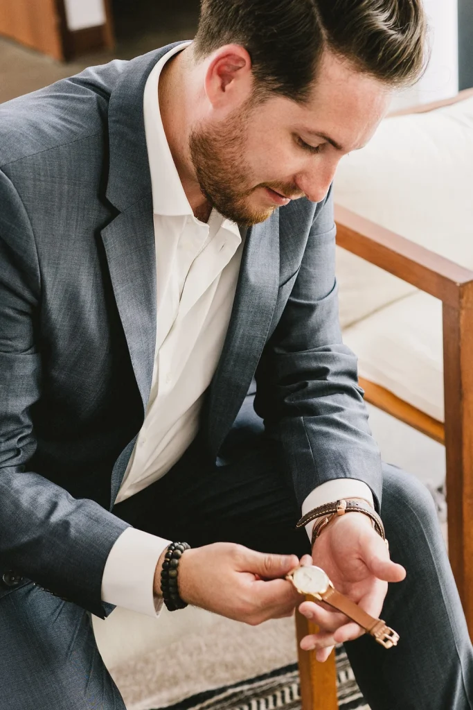 Groom in a navy suit checking his watch while sitting in a modern setting.