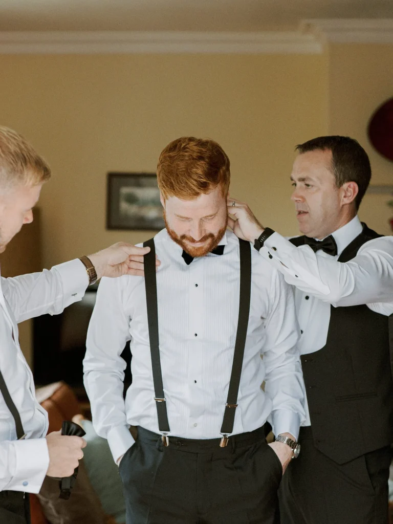 A groom-to-be with red hair smiles as his groomsmen adjust his suspenders and bow tie.