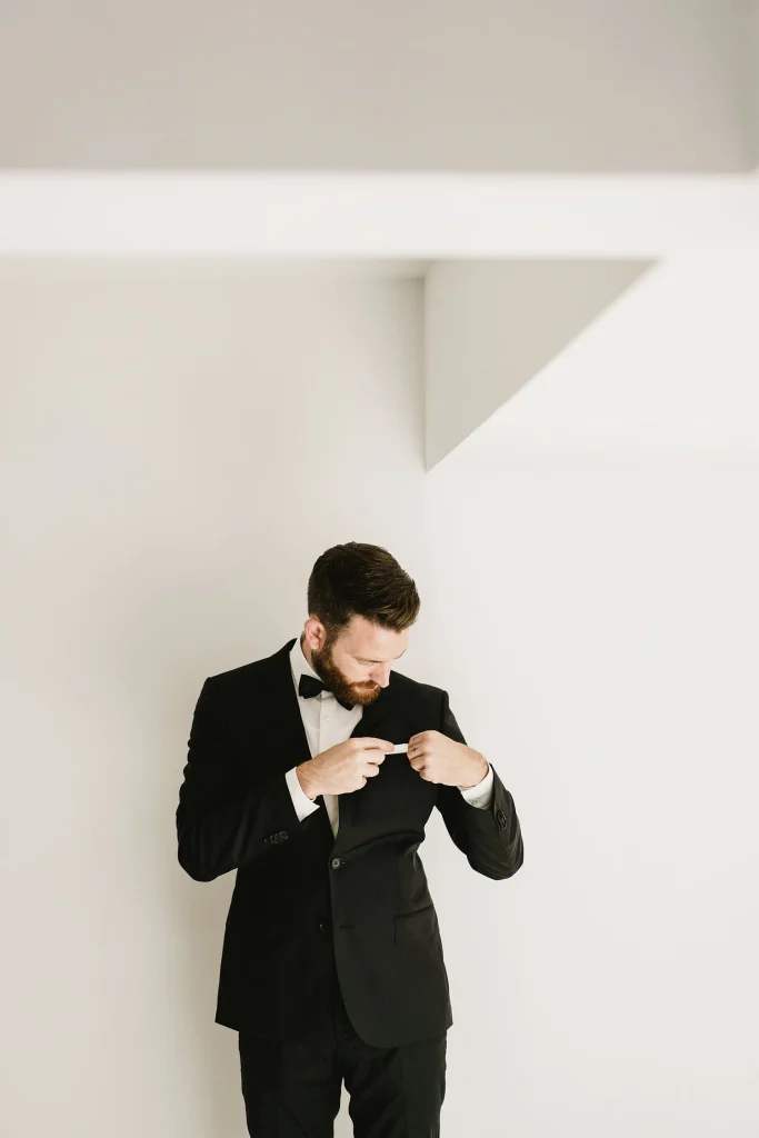 A groom in a black tuxedo adjusts his jacket, standing against a minimalist white wall.