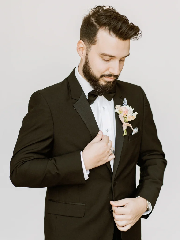 A groom in a black tuxedo and bowtie adjusts his suit jacket while looking down, with a soft, neutral background highlighting his boutonniere.