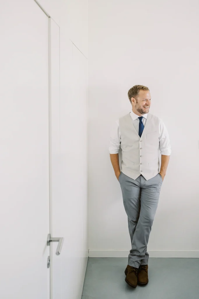 Groom in a light grey vest and navy tie leaning casually against a white wall, smiling.