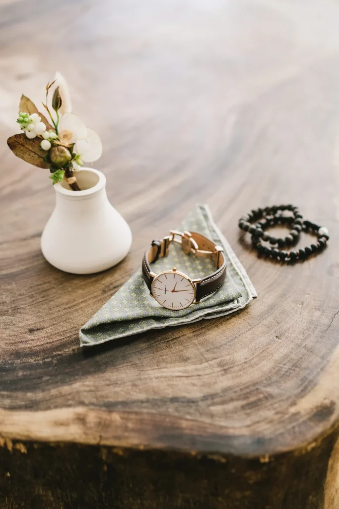 Groom's wedding accessories including a leather strap watch, a boutonnière, and a beaded bracelet on a wooden table.