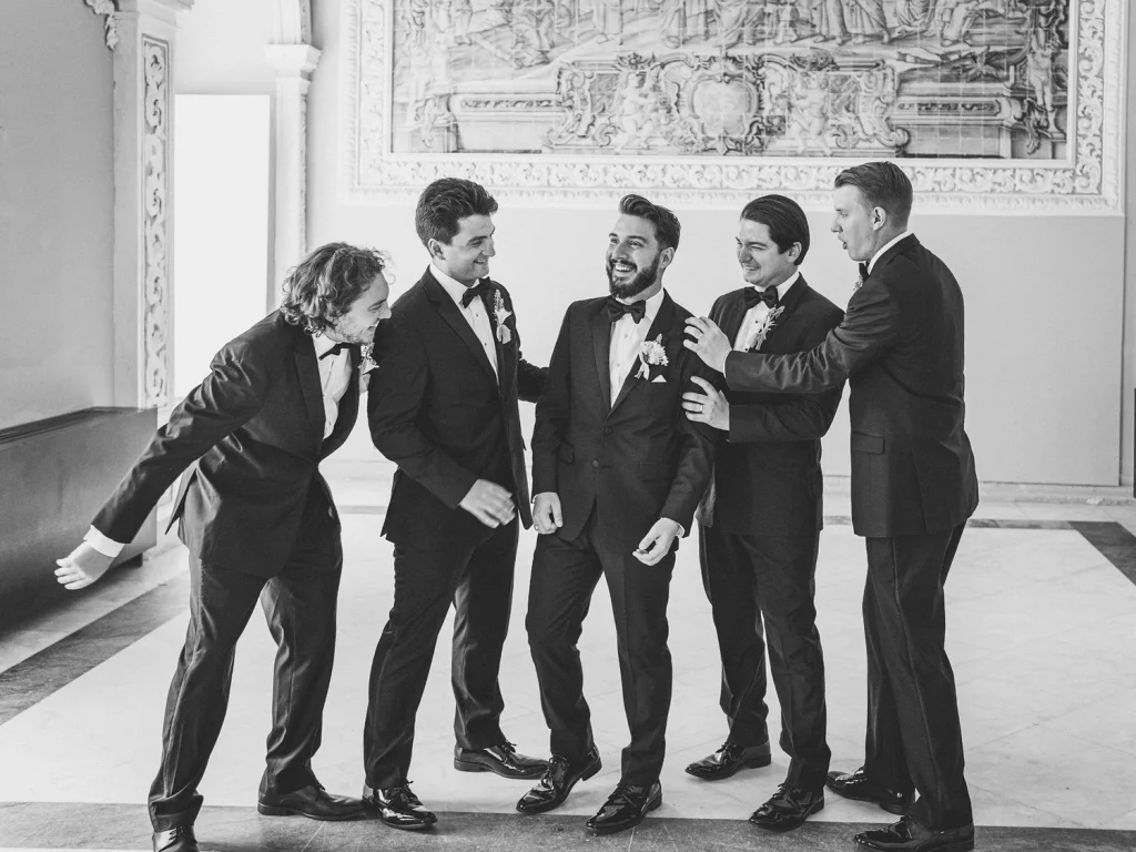 A group of groomsmen in classic black tuxedos shares a candid moment, laughing together in an elegant venue.