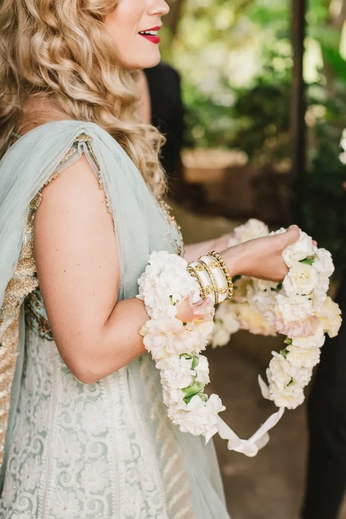 A wedding guest dressed in an elegant pastel green and gold outfit holds a delicate floral garland at Quinta de Sant’Ana, Portugal. The intricate embroidery, gold bangles, and soft curls add to the festive and cultural ambiance.