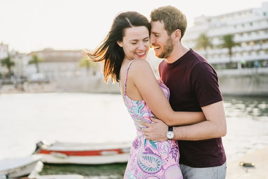 A couple embraces near the water, laughing as the wind blows through the woman's hair, with a red boat in the background.