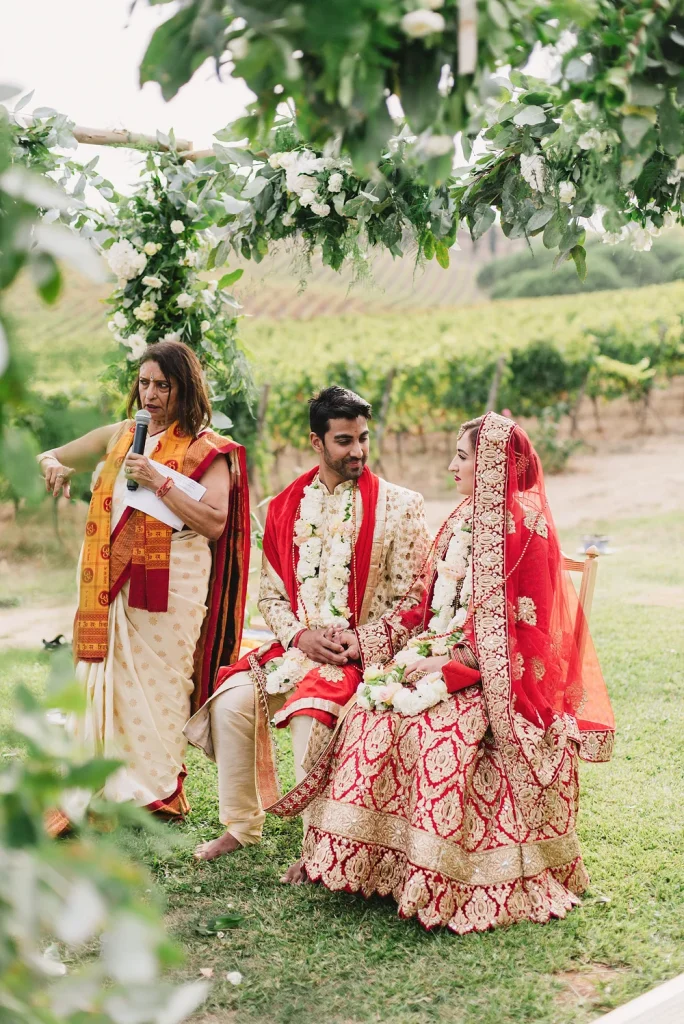 A bride and groom sit under a floral mandap during their Hindu wedding ceremony at Quinta de Sant’Ana, Portugal. Dressed in traditional red and gold attire, they listen to the priest as rolling vineyards provide a breathtaking natural backdrop.