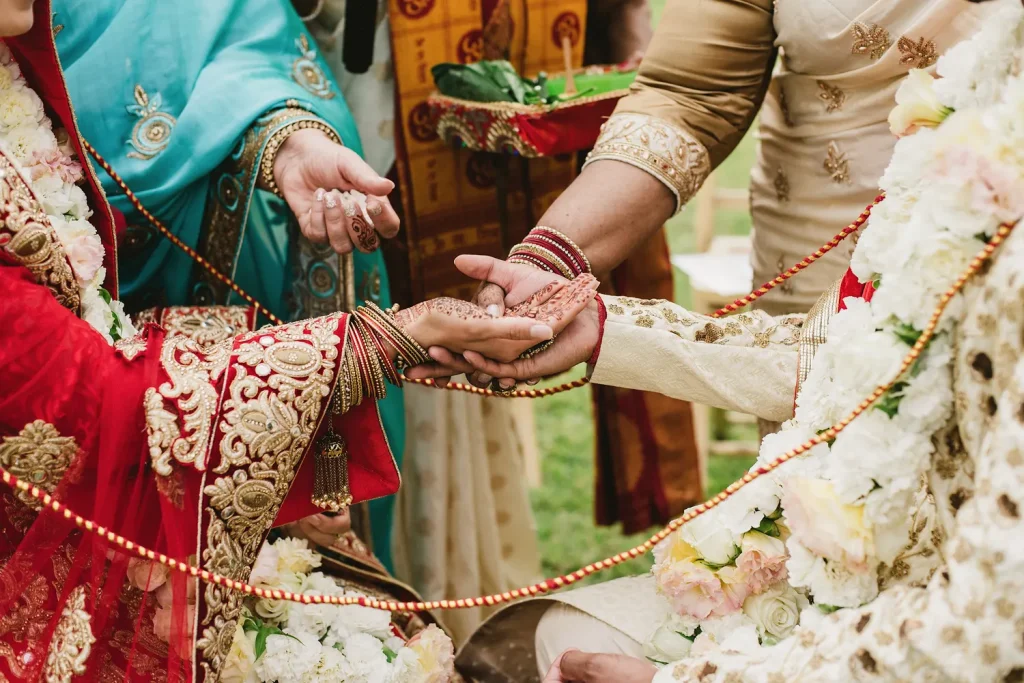 A close-up of a sacred Hindu wedding ritual at Quinta de Sant’Ana, Portugal. The bride and groom’s henna-adorned hands are joined together by family members as part of the ceremony, surrounded by intricate red and gold attire, floral garlands, and symbolic offerings.