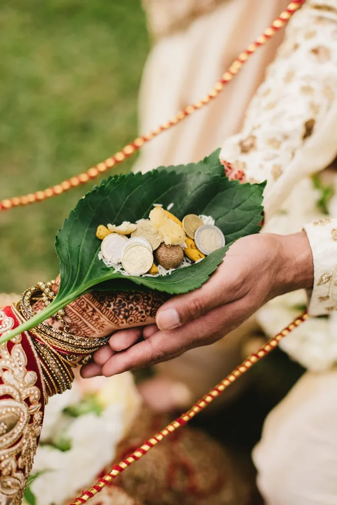 A close-up of a Hindu wedding ritual offering at Quinta de Sant’Ana, Portugal. The groom’s and bride’s hands, adorned with henna and gold jewelry, hold a green leaf containing coins, rice, and symbolic offerings, representing prosperity and blessings.