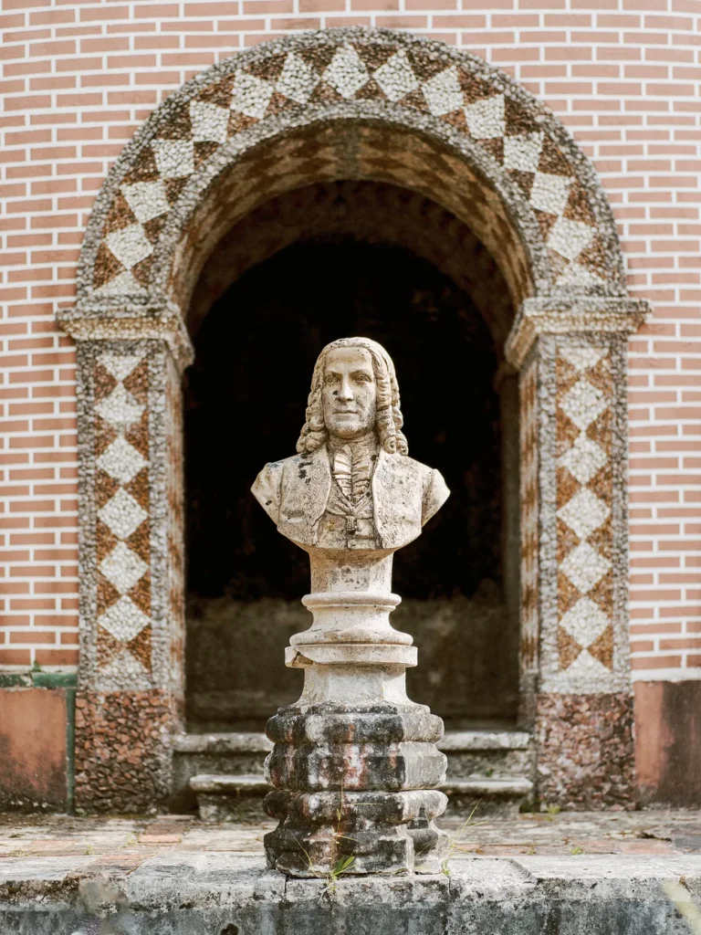 A weathered stone bust of a historical figure stands in front of an arched brick entryway adorned with geometric patterns.