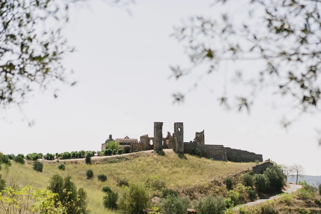 Ancient stone ruins on a hilltop in Portugal, framed by tree branches, with a winding road leading up to the site.