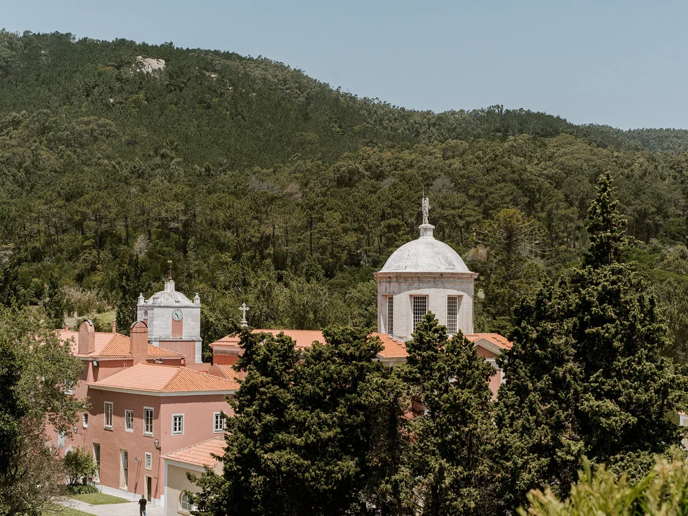 A historic monastery with terracotta rooftops and domed towers nestled against a lush green mountain landscape under a clear blue sky.