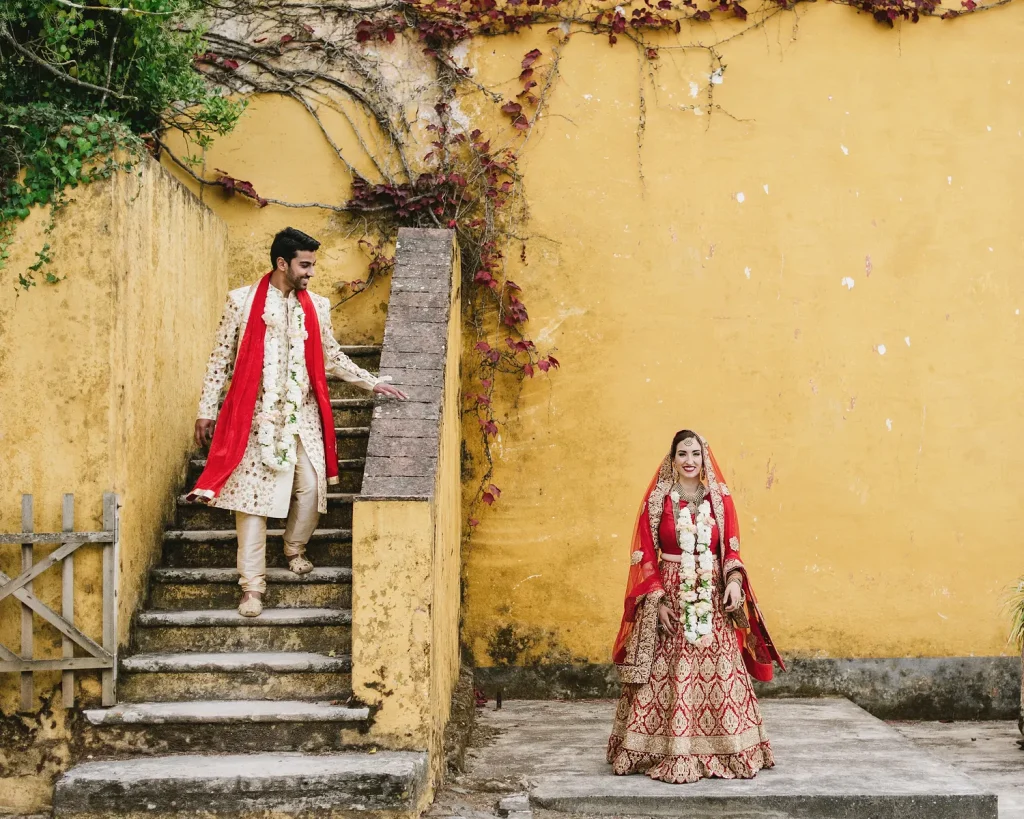 Bride and groom in traditional Indian wedding attire posing against a vibrant yellow wall with vines.
