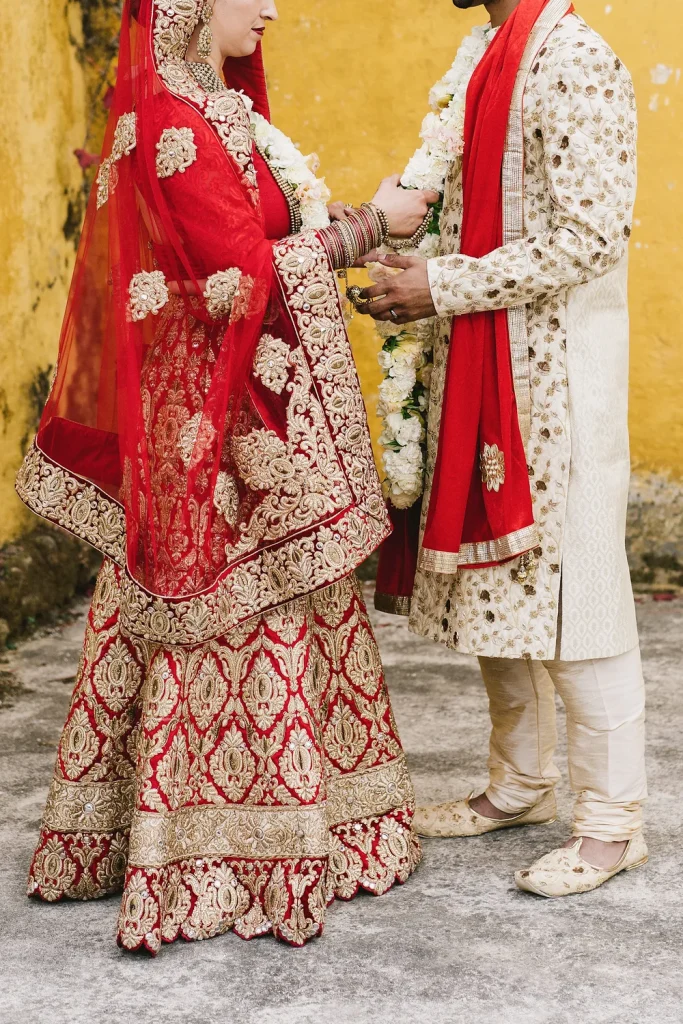 A bride and groom in traditional Indian wedding attire exchange floral garlands at Quinta de Sant’Ana, Portugal. The bride wears a red and gold embroidered lehenga with intricate jewelry, while the groom is dressed in a cream sherwani with gold detailing and a matching red stole. The vibrant yellow wall in the background enhances the rich colors of their outfits, creating a striking and elegant scene.