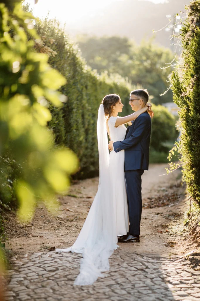 Bride and groom standing close together, bathed in warm sunlight, with the bride’s veil flowing behind her.