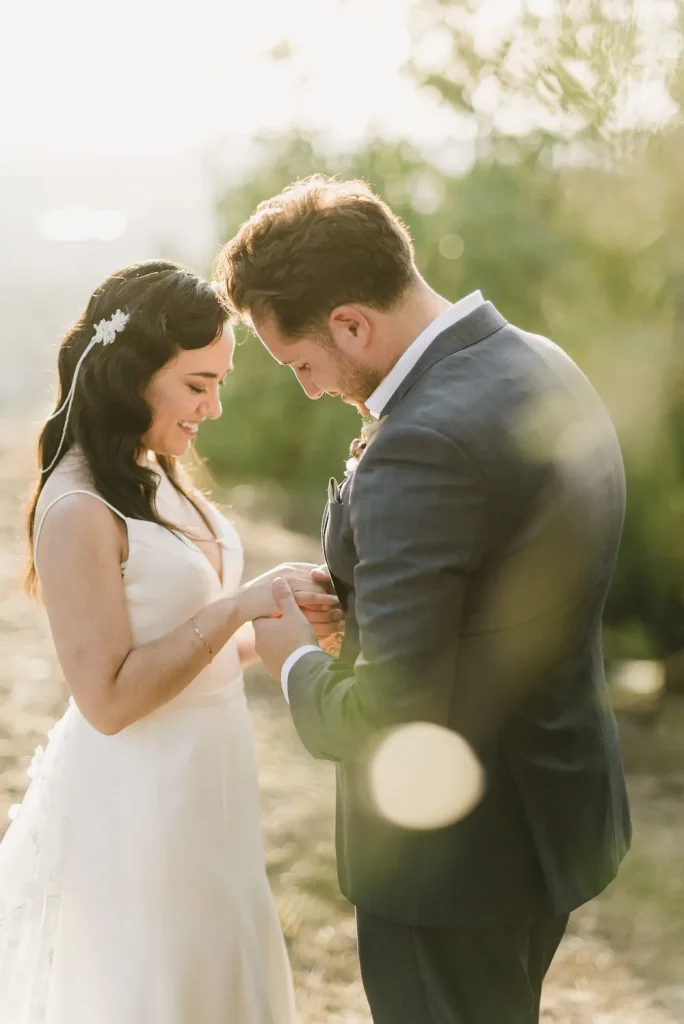 Bride and groom exchanging rings in a sunlit outdoor setting, smiling as they share a private moment.