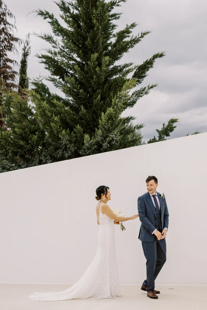 A bride in a sleek white gown surprises her groom during their first look moment. The groom, in a navy suit, reacts with excitement against a modern white wall with tall green trees in the background.