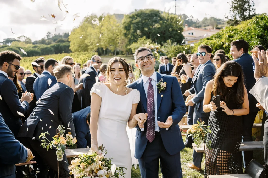 A newlywed couple walks down the aisle, laughing as guests toss flower petals in celebration during an outdoor vineyard wedding, surrounded by friends and family dressed in elegant attire.