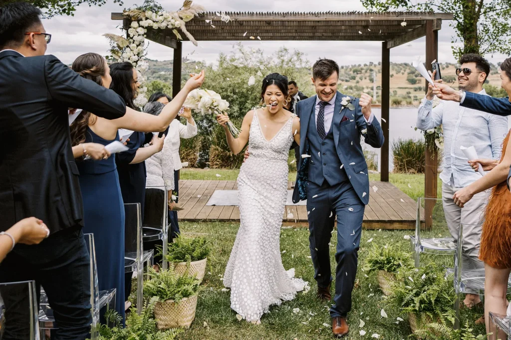 A joyful newlywed couple walks down the aisle as guests toss flower petals in celebration, with a floral-adorned pergola and scenic lake in the background.