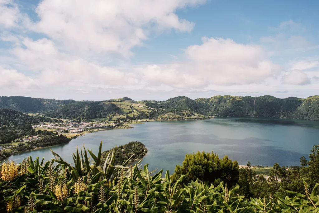 A breathtaking panoramic view of a deep blue lake surrounded by rolling green mountains and a small village, framed by tropical plants in the foreground under a bright blue sky with scattered clouds.