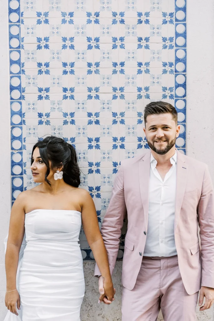 A stylish couple poses in front of a traditional Portuguese blue and white tiled wall, holding hands and smiling.