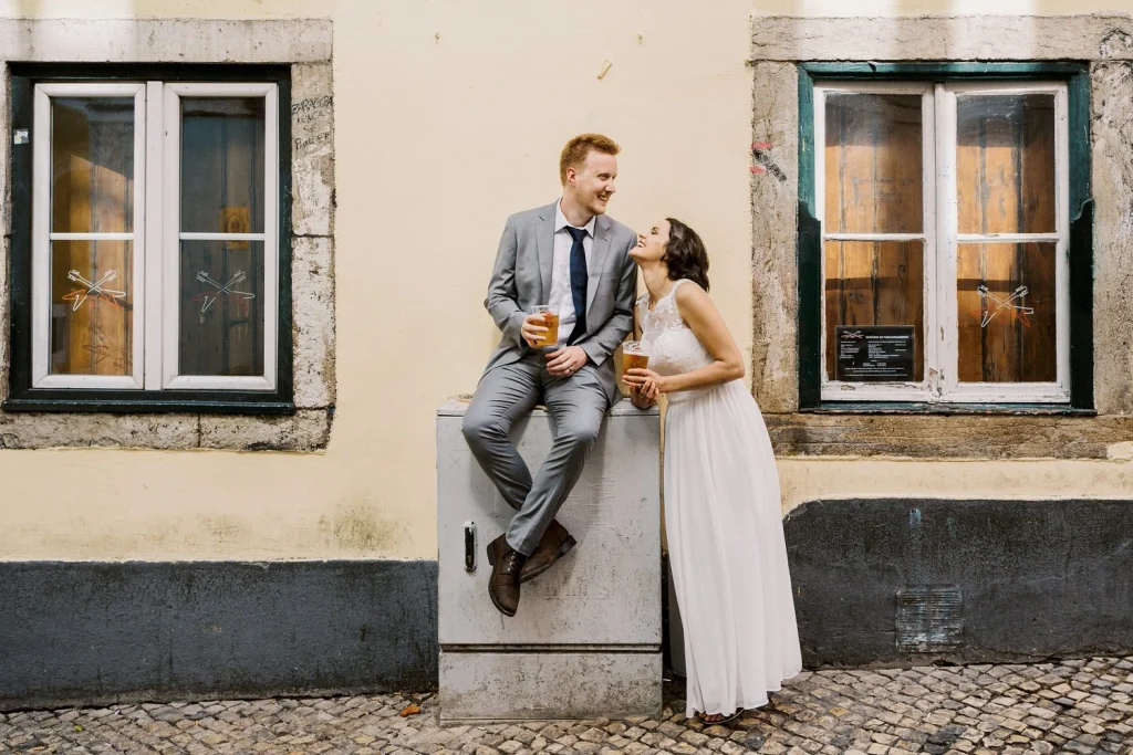A groom sits on a ledge sipping a drink while his bride leans in with a loving gaze, against a European-style building.