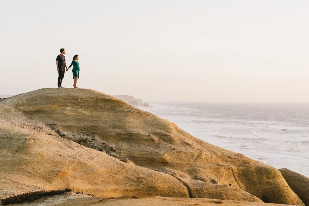 Pregnant woman in a flowing teal dress holding hands with her partner on a scenic coastal rock formation.
