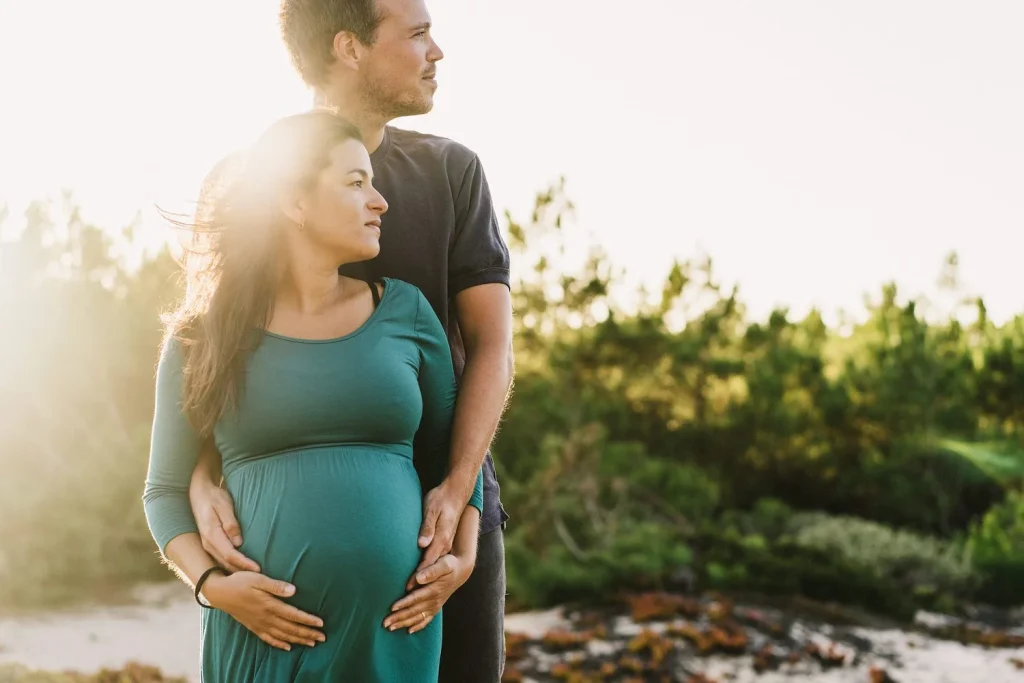 Expecting couple standing together, with the man's arms wrapped around the woman's pregnant belly in golden sunlight.