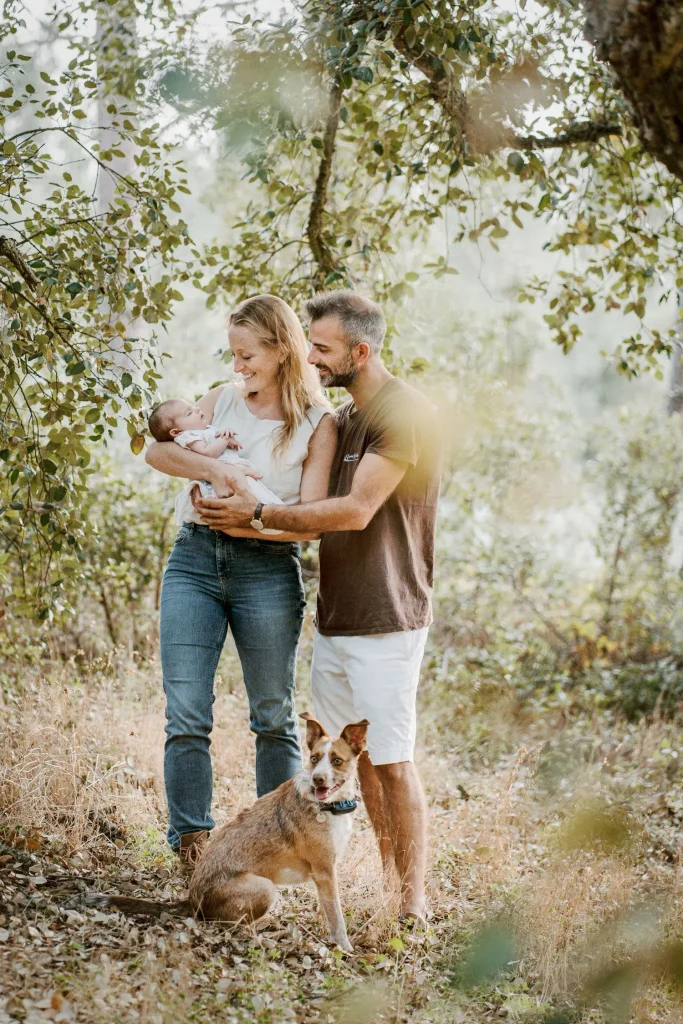 A young family with their newborn and pet dog, standing in a sunlit natural setting.