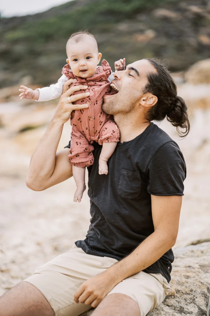 Father playfully lifting his baby in the air, pretending to take a bite, with a beach backdrop.